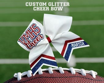 May include: A white cheer bow with red and blue glitter accents, resting on a football. The bow features the text "VMHS Cheer" and "MARYZA". The top of the image reads "CUSTOM GLITTER CHEER BOW".