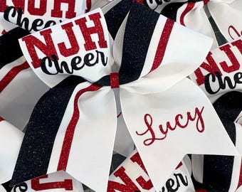 May include: White cheer bow with black and red glitter stripes. The bow has the text "NJH Cheer" in red glitter and the name "Lucy" in red glitter.