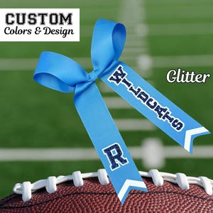 May include: A blue glitter cheer bow with the words "Wildcats" and "R" in navy blue glitter. The bow is on top of a brown football.