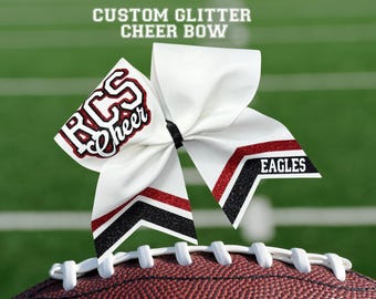 May include: A white cheer bow with red and black glitter accents, resting on a football. The bow features the text "RCS Cheer" and "EAGLES". The top of the image reads "CUSTOM GLITTER CHEER BOW".