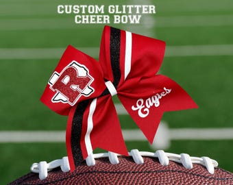 May include: A red cheer bow with black and white stripes, and the word "Eagles" in white script. The bow has a glittery "R" logo. The text "CUSTOM GLITTER CHEER BOW" is at the top. The bow is resting on a football.