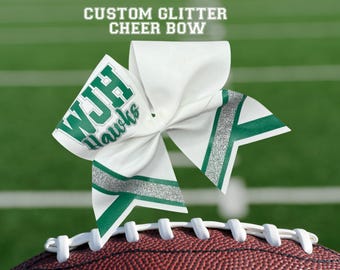 May include: A white cheer bow with green and silver accents, resting on a football. The bow features the text "WJH Hawks" in green. The words "CUSTOM GLITTER CHEER BOW" are at the top of the image.