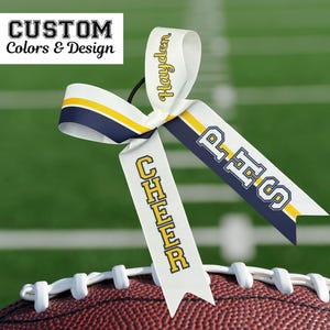 Puede incluir: Un lazo de animadora blanco con detalles en azul marino y amarillo, personalizado con el nombre "Hayden" y las letras "PHS". El lazo est&aacute; colocado sobre un bal&oacute;n de f&uacute;tbol americano. El texto en la parte superior dice "CUSTOM Colors & Design".