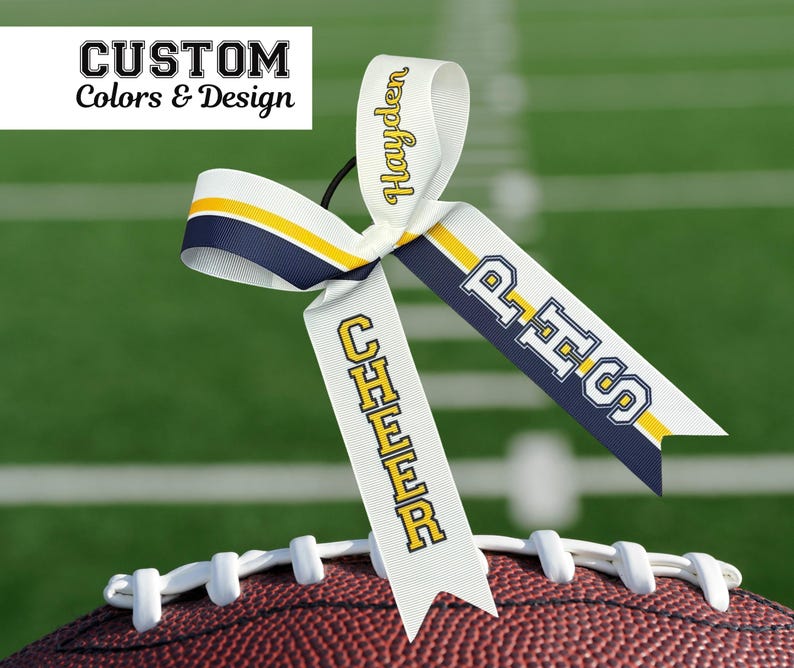 Puede incluir: Un lazo de animadora blanco con detalles en azul marino y amarillo, personalizado con el nombre "Hayden" y las letras "PHS". El lazo est&aacute; colocado sobre un bal&oacute;n de f&uacute;tbol americano. El texto en la parte superior dice "CUSTOM Colors & Design".