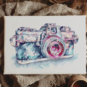 May include: Cross-stitch art of a vintage camera in shades of blue, pink, and purple. The camera is on a white canvas, with a detailed lens and body. The background includes a rustic wooden surface and fabric.