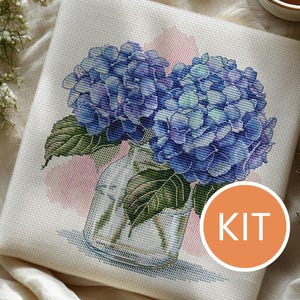 Può includere: Kit per punto croce con un'illustrazione dettagliata di fiori di ortensia blu in un vaso di vetro. I fiori hanno sfumature di blu e viola, con foglie verdi. La parola "KIT" è in un cerchio arancione.