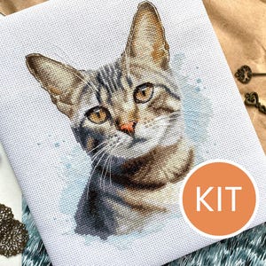 Può includere: Kit per punto croce con un ritratto dettagliato di un gatto tigrato con pelo marrone e grigio, su sfondo acquerello azzurro. Il kit include la parola "KIT" in un cerchio arancione, insieme a filo da ricamo ed elementi decorativi.