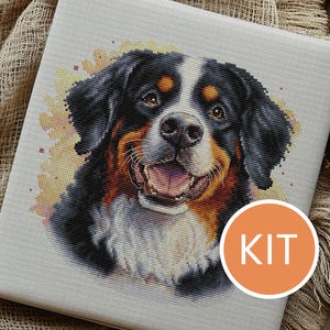 Può includere: Kit per punto croce raffigurante un Bovaro del Bernese. Il muso del cane è dettagliato con filo nero, marrone e bianco, con la bocca aperta e un'espressione amichevole. La parola "KIT" è in un cerchio arancione.