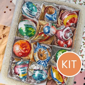 Può includere: Un kit per punto croce con una scatola di ornamenti natalizi colorati. Gli ornamenti sono di varie forme e colori, tra cui rosso, verde, blu e giallo. La parola "KIT" è in un cerchio in basso a destra.