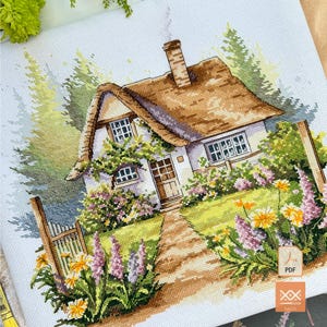 Könnte beinhalten: Ein Kreuzstichmuster auf hellblauem Stoff, das ein Häuschen mit Reetdach, einem Steinschornstein und einer Holztür zeigt. Die Szene umfasst einen Gartenweg, ein Holztor und bunte Blumen in den Farben Gelb, Violett und Grün.