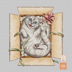 May include: A cross-stitch pattern featuring a light gray cat with blue eyes lying on its back inside an open cardboard box. The cat is surrounded by white packing peanuts and green ribbon, with a large pink bow near the top right corner.