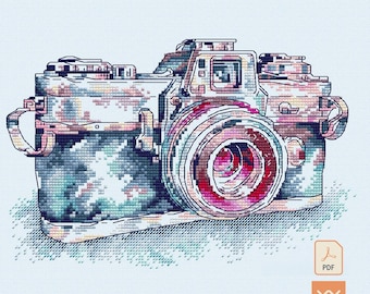 May include: A cross-stitch pattern of a camera featuring a pixelated design in shades of blue, pink, and white. The camera body has a textured appearance with a prominent lens, strap attachments on the sides, and a small PDF icon in the corner.
