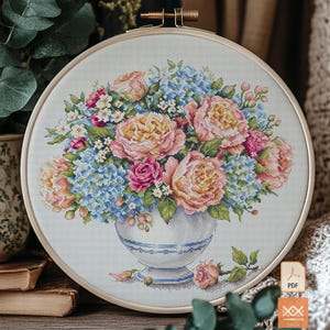 May include: A circular wooden embroidery hoop containing a cross-stitch pattern of a floral bouquet in a white vase with blue trim. The bouquet features pink and yellow peonies, blue hydrangeas, and small white flowers with green leaves.