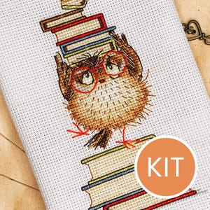 Può includere: Kit per punto croce con un gufo marrone con occhiali rossi, che trasporta pile di libri. Il gufo è ricamato su tessuto bianco. Un adesivo arancione con la scritta "KIT" è nell'angolo. Una chiave e una fetta d'arancia secca sono sullo sfondo.