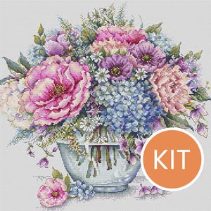 Può includere: Un disegno floreale a punto croce con un bouquet di peonie rosa, ortensie blu e margherite bianche in un vaso di vetro. L'immagine ha un aspetto morbido, simile a un acquerello. Un cerchio arancione con la parola "KIT" è nell'angolo in basso a destra.