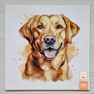 May include: A cross-stitch pattern featuring a golden Labrador retriever dog with a happy expression, open mouth, and pink tongue. The design is set against a light background with soft yellow watercolor-style accents and includes a PDF icon.