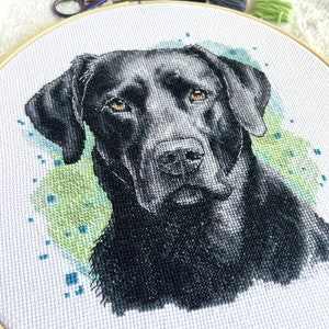 Puede incluir: Bordado de punto de cruz de la cabeza de un labrador retriever negro con un fondo de estilo acuarela en verde y azul.