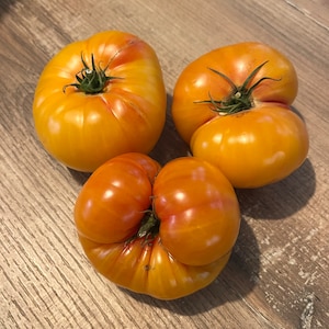 Op de afbeelding: Drie grote, rijpe, gele tomaten met rode strepen. De tomaten liggen op een houten oppervlak.