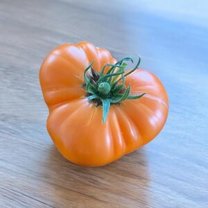 Puede incluir: Un tomate grande, naranja, de herencia con un tallo y hojas verdes. El tomate tiene forma de corazón y una textura ligeramente irregular.