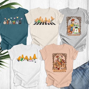 May include: Five different t-shirts with various cartoon characters and designs. The shirts are all white, light beige, light blue, and light pink. The designs include a group of cartoon characters walking across a crosswalk, a group of cartoon characters wearing sombreros, a cartoon character riding a horse, and a cartoon character with a friend.