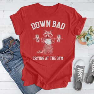 May include: Red t-shirt with a white graphic of a raccoon lifting weights and the text "Down Bad Crying at the Gym".