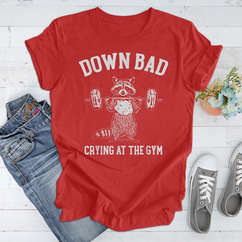May include: Red t-shirt with a white graphic of a raccoon lifting weights and the text "Down Bad Crying at the Gym".