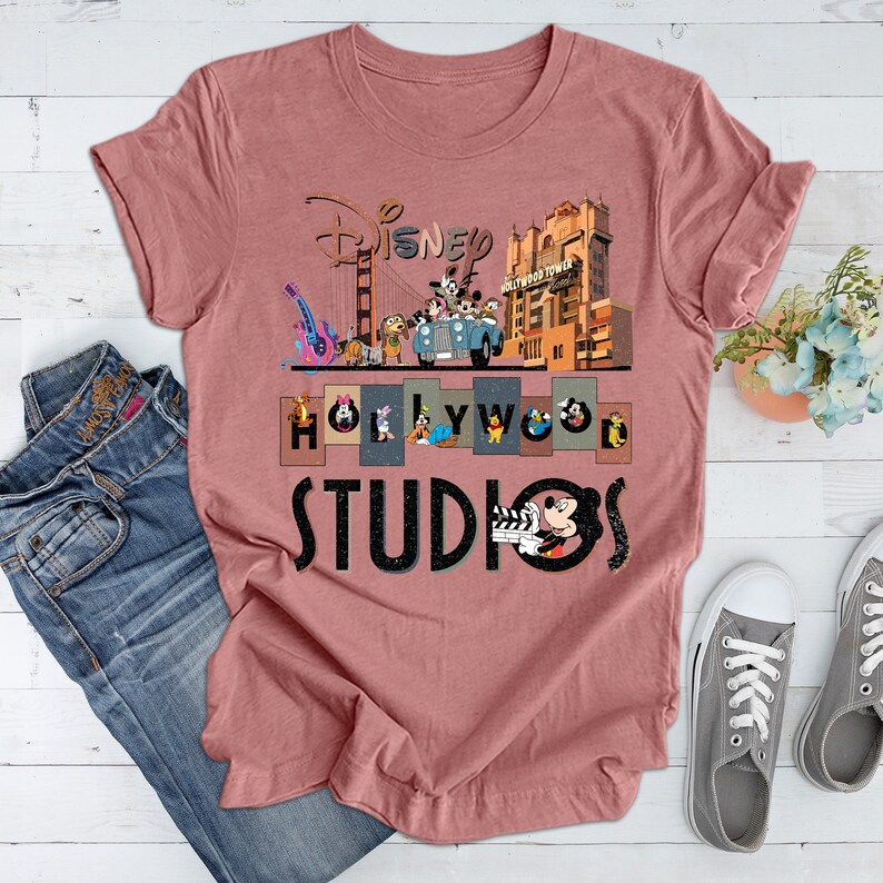 May include: A pink t-shirt with a graphic print of Disney characters in front of the Hollywood Tower Hotel and the Golden Gate Bridge. The text "Disney Hollywood Studios" is printed below the graphic.