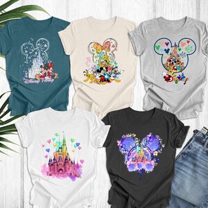 May include: Five different t-shirts featuring various Disney characters and designs. The shirts are all white, grey, black, and teal. The designs include Mickey Mouse, Minnie Mouse, Donald Duck, Goofy, Pluto, and Cinderella's castle. The shirts are all printed with the word "Disney" on them.