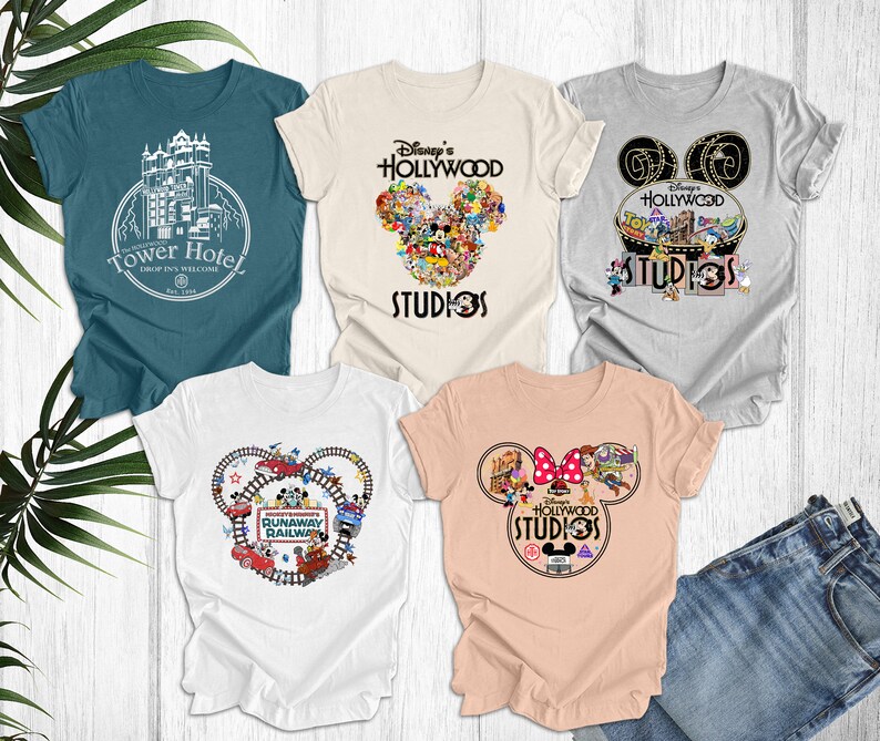 May include: Five different t-shirts featuring Disney's Hollywood Studios. The shirts are white, light blue, gray, and peach. The designs include Mickey Mouse ears, the Tower of Terror, and the Runaway Railway.