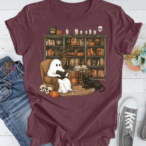 May include: Burgundy t-shirt featuring a Halloween-themed illustration. A ghost reads in a chair in front of a bookshelf, with pumpkins, candles, and a black cat. Skulls and pumpkins are on the floor. The design is printed on a burgundy t-shirt.
