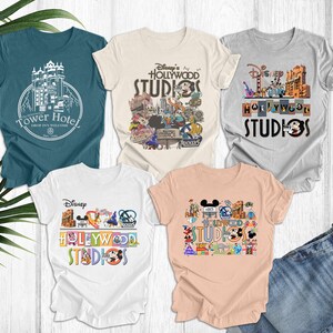 May include: Four different t-shirts featuring Disney's Hollywood Studios. The shirts are in various colors, including teal, beige, gray, and peach. Each shirt features a different design, including the Tower of Terror, Mickey Mouse, and other Disney characters.