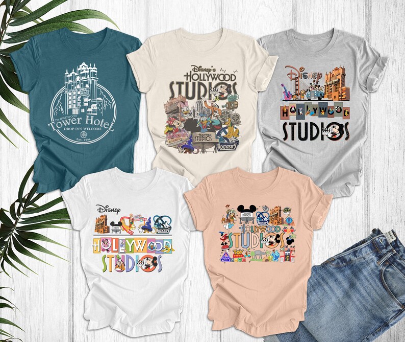 May include: Four different t-shirts featuring Disney's Hollywood Studios. The shirts are in various colors, including teal, beige, gray, and peach. Each shirt features a different design, including the Tower of Terror, Mickey Mouse, and other Disney characters.