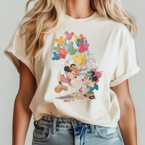 May include: A white t-shirt with a graphic print of Mickey Mouse and Minnie Mouse holding balloons in front of Cinderella's Castle. The balloons are in various colors, including red, yellow, blue, and green.