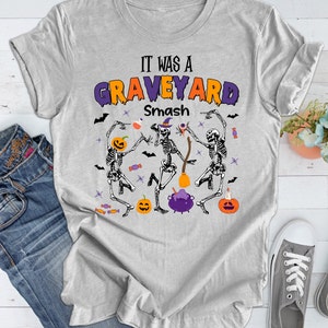 May include: Gray t-shirt with the text "It was a Graveyard Smash" in purple and orange letters. Three skeletons are dancing with a cauldron, bats, and candy.