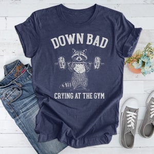 May include: A navy blue t-shirt with a white graphic of a raccoon lifting weights. The text "Down Bad Crying At The Gym" is printed below the raccoon.