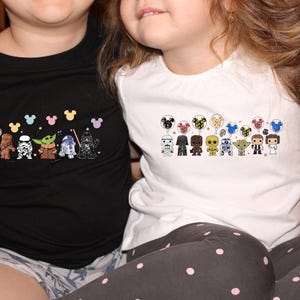 May include: Two children wearing black and white t-shirts with Star Wars characters and Mickey Mouse ears. The shirts feature a line of Star Wars characters holding balloons with Mickey Mouse ears.