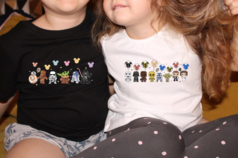 May include: Two children wearing black and white t-shirts with Star Wars characters and Mickey Mouse ears. The shirts feature a line of Star Wars characters holding balloons with Mickey Mouse ears.