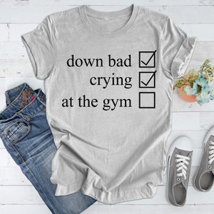 May include: A gray t-shirt with a checklist that reads "down bad", "crying", and "at the gym". The first two items are checked off, while the last item is unchecked.