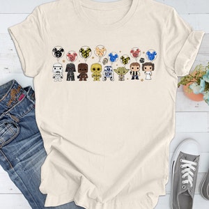 May include: A beige t-shirt with a graphic print of Star Wars characters holding Mickey Mouse balloons. The characters include Darth Vader, Chewbacca, R2-D2, Yoda, and others.