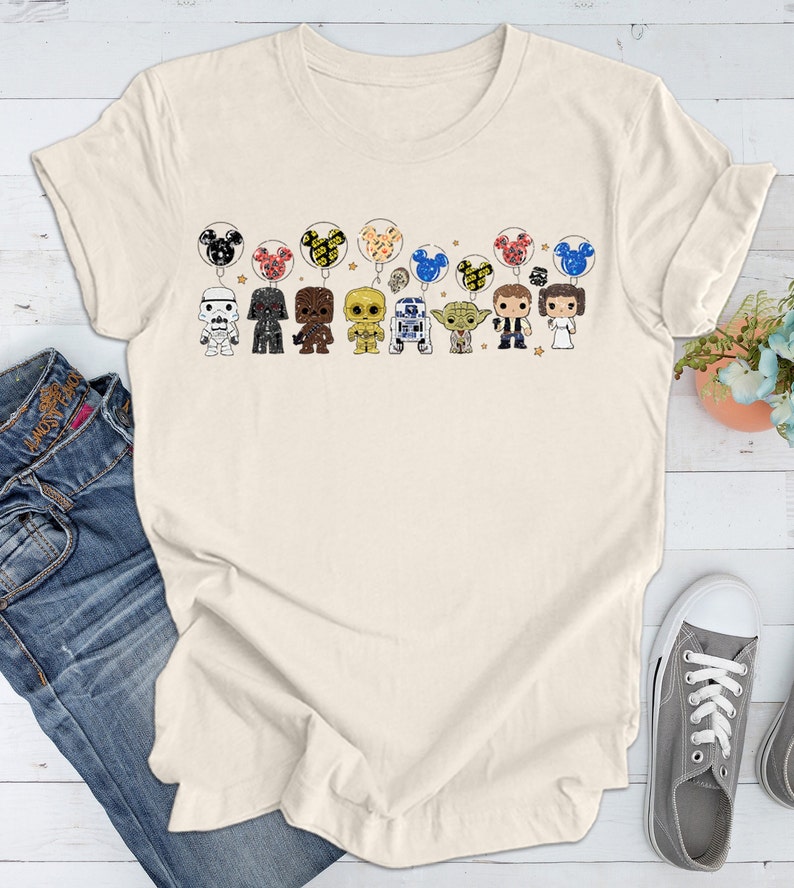 May include: A beige t-shirt with a graphic print of Star Wars characters holding Mickey Mouse balloons. The characters include Darth Vader, Chewbacca, R2-D2, Yoda, and others.