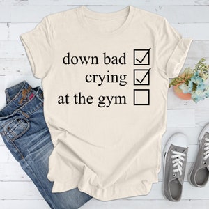 May include: A white t-shirt with a black checklist that reads "down bad", "crying", and "at the gym". The first two options are checked, while the last option is unchecked.