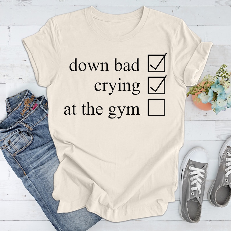 May include: A white t-shirt with a black checklist that reads "down bad", "crying", and "at the gym". The first two options are checked, while the last option is unchecked.