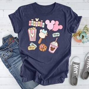 May include: A navy blue t-shirt with a whimsical design featuring Mickey Mouse ears, popcorn, a Mickey Mouse ice cream bar, a Mickey Mouse cookie, a Mickey Mouse milkshake, and the word "Magical" in pink letters.