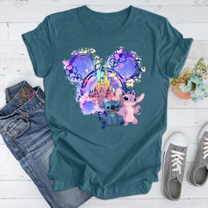 Puede incluir: Camiseta de color turquesa con un gr&aacute;fico colorido de Stitch y Angel, un castillo y orejas de Mickey Mouse. La camiseta se combina con vaqueros azules y zapatillas grises, creando un look casual.