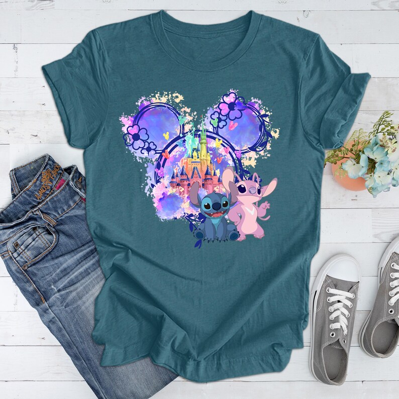 Puede incluir: Camiseta de color turquesa con un gr&aacute;fico colorido de Stitch y Angel, un castillo y orejas de Mickey Mouse. La camiseta se combina con vaqueros azules y zapatillas grises, creando un look casual.