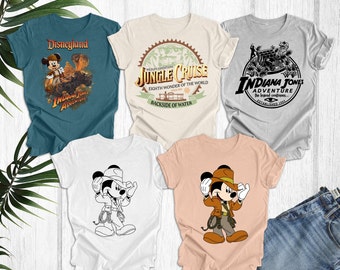 May include: Five different t-shirts featuring various Disney characters and attractions. The shirts are in different colors, including teal, beige, gray, white, and peach. The designs include Mickey Mouse dressed as Indiana Jones, the Jungle Cruise logo, the Indiana Jones Adventure logo, and a black and white line drawing of Mickey Mouse dressed as Indiana Jones.