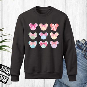 Puede incluir: Sudadera negra de cuello redondo con un estampado de nueve dise&ntilde;os coloridos en forma de rat&oacute;n con lazos. Los lazos son de color rosa, azul, amarillo y verde. La sudadera se muestra con vaqueros azules y zapatillas deportivas blancas y negras.