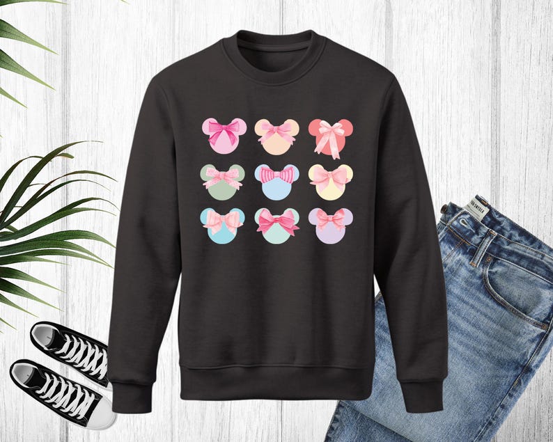 Puede incluir: Sudadera negra de cuello redondo con un estampado de nueve dise&ntilde;os coloridos en forma de rat&oacute;n con lazos. Los lazos son de color rosa, azul, amarillo y verde. La sudadera se muestra con vaqueros azules y zapatillas deportivas blancas y negras.