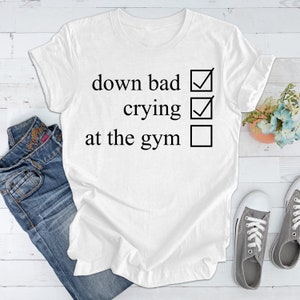 May include: White t-shirt with black text that reads "down bad", "crying", and "at the gym" with checkboxes next to each phrase. The first two checkboxes are checked, and the last one is unchecked.