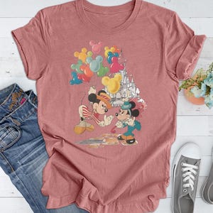 May include: A pink t-shirt with a vintage-style graphic of Mickey Mouse and Minnie Mouse holding balloons in front of a castle. The graphic is in shades of red, yellow, blue, and green.